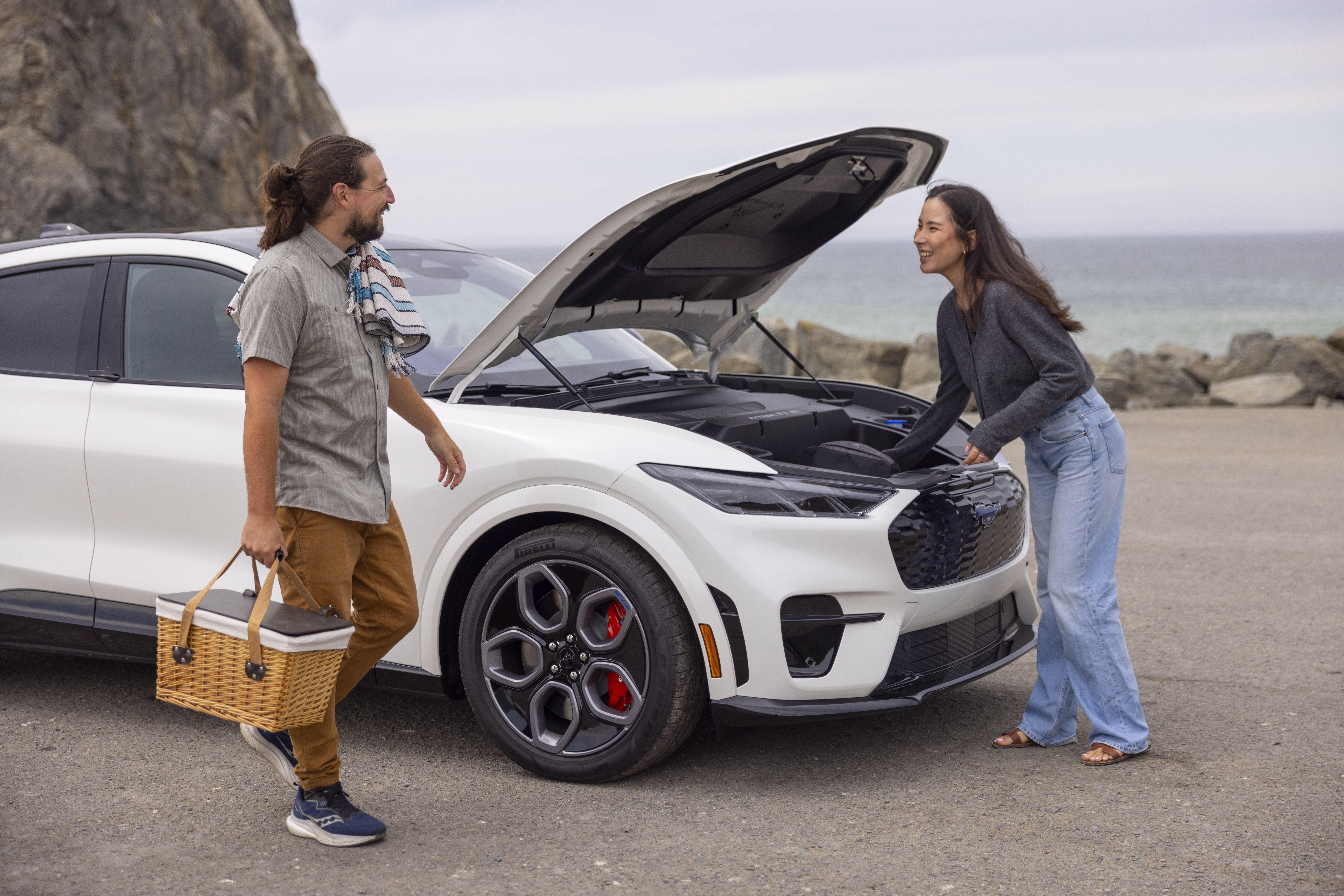 A man and woman stand on the beach holding a picnic basket near a white Mustang Mach-E.